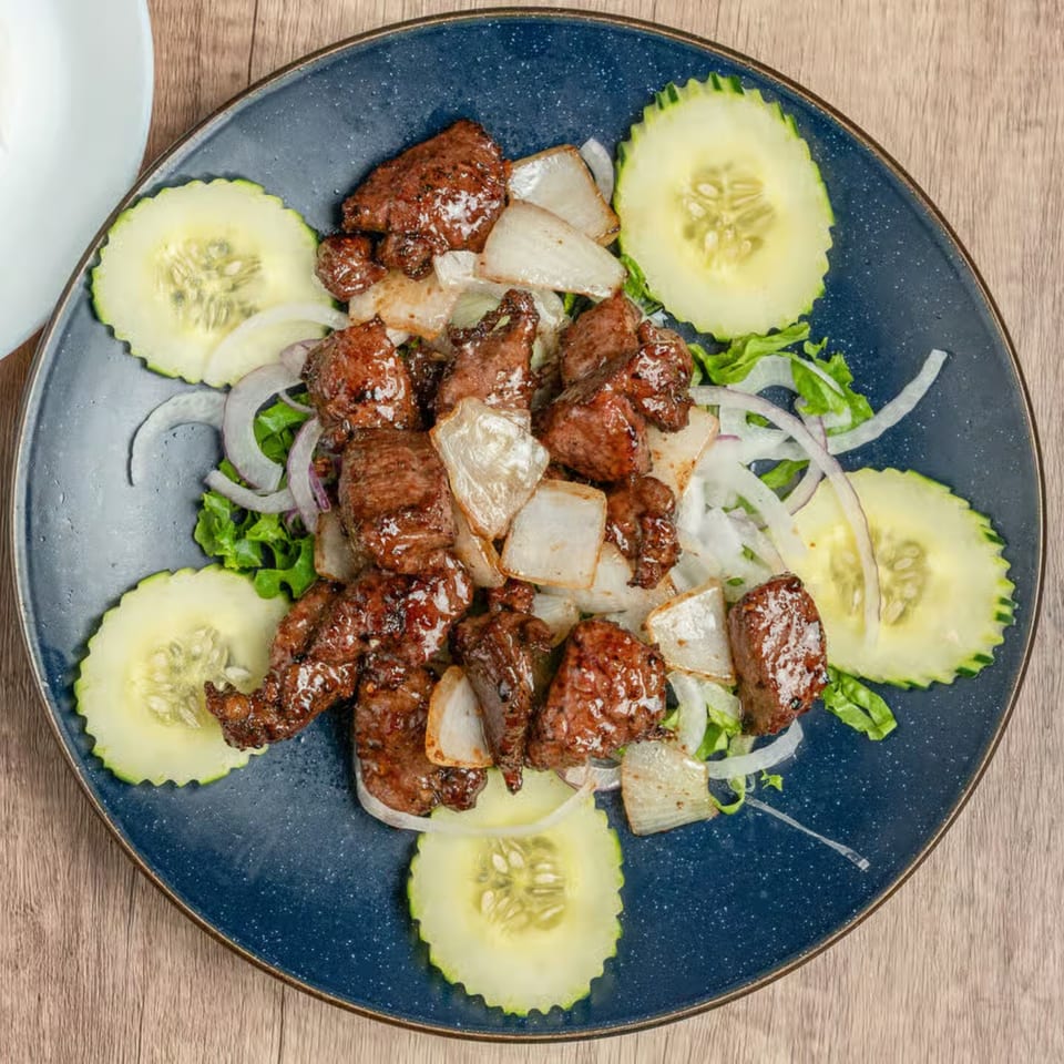Vietnamese shaking beef (Bo Luc Lac) with onions, lettuce, and cucumber slices on a blue plate.
