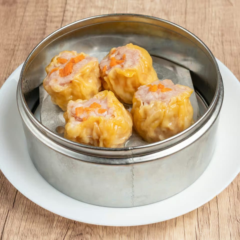 Four steamed pork and shrimp shumai dumplings with orange garnish in a metal steamer basket.